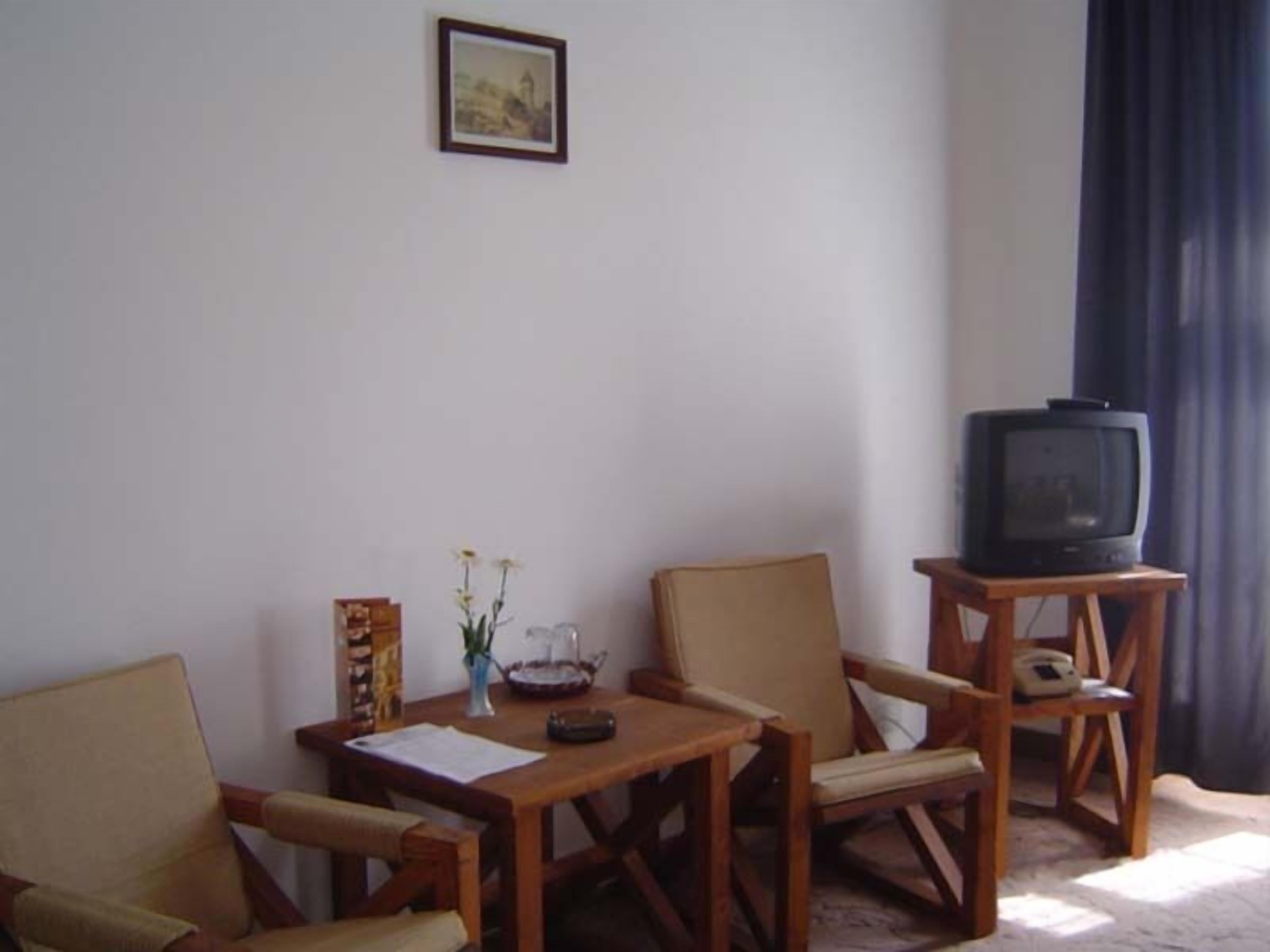 A room with two wooden chairs, a small table with a vase of flowers, a framed picture on the wall, and a television on a wooden stand.