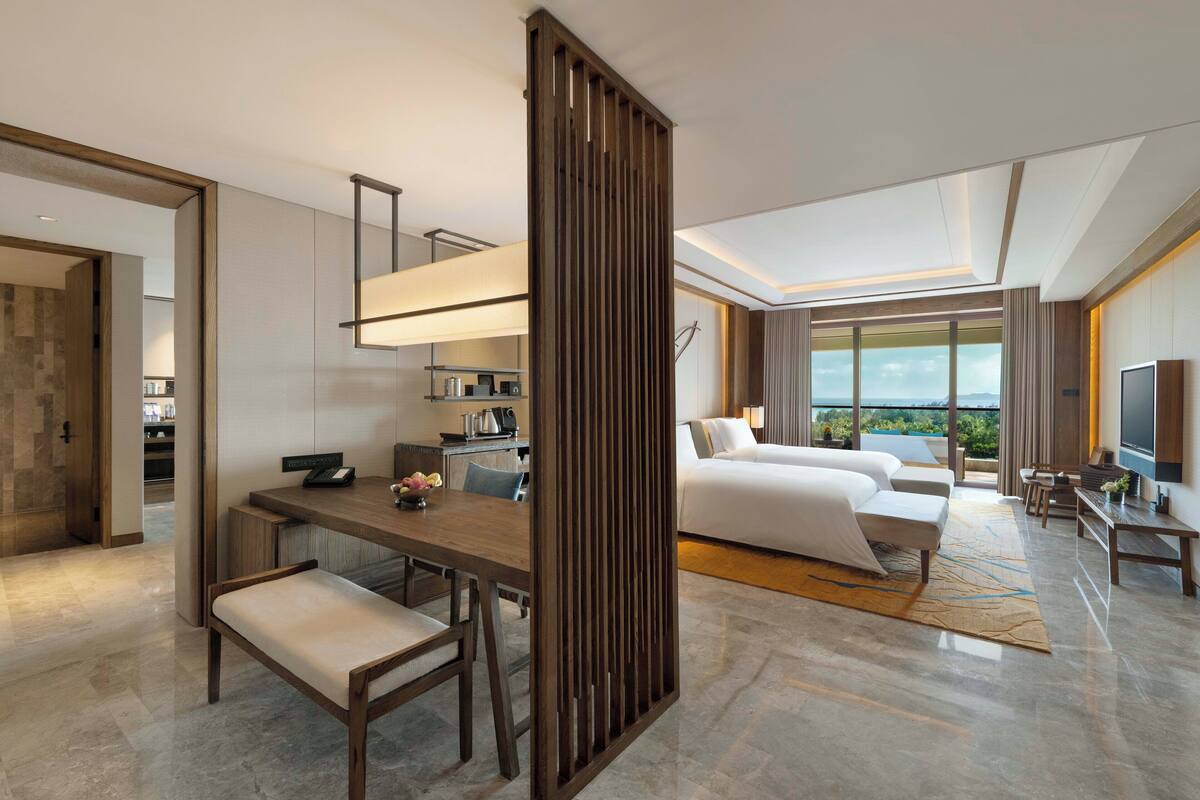 A modern hotel room with a dining area, a large bed, a TV, and a balcony with a view.