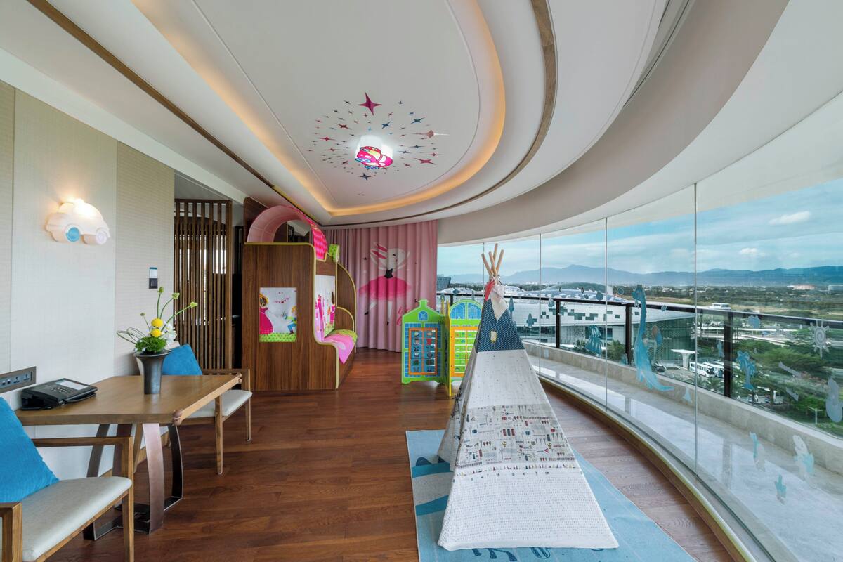 A modern hotel room with a panoramic city view, a wooden floor, a dining area with a table and chairs, and a children's play area with colorful furniture.