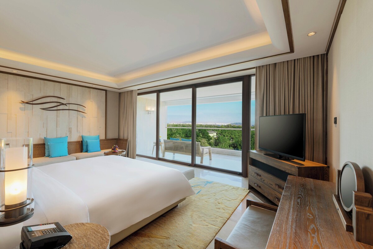 Ein Hotelzimmer mit einem großen Bett, einem Fernseher, einem Schreibtisch und einem Balkon mit Blick.