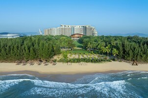 A beachfront hotel with a large building, a sandy beach, and a forested area.