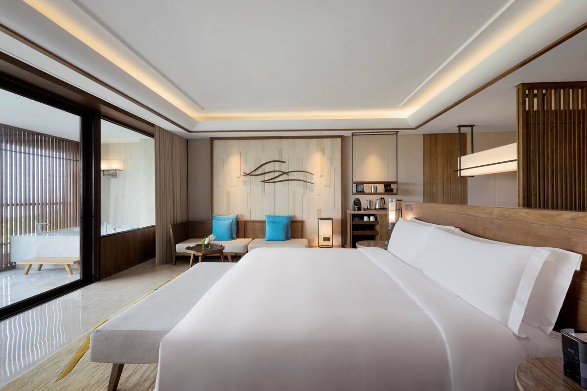 A modern hotel room with a large bed, a seating area with a sofa and chairs, a small table, and a view of the outside balcony.