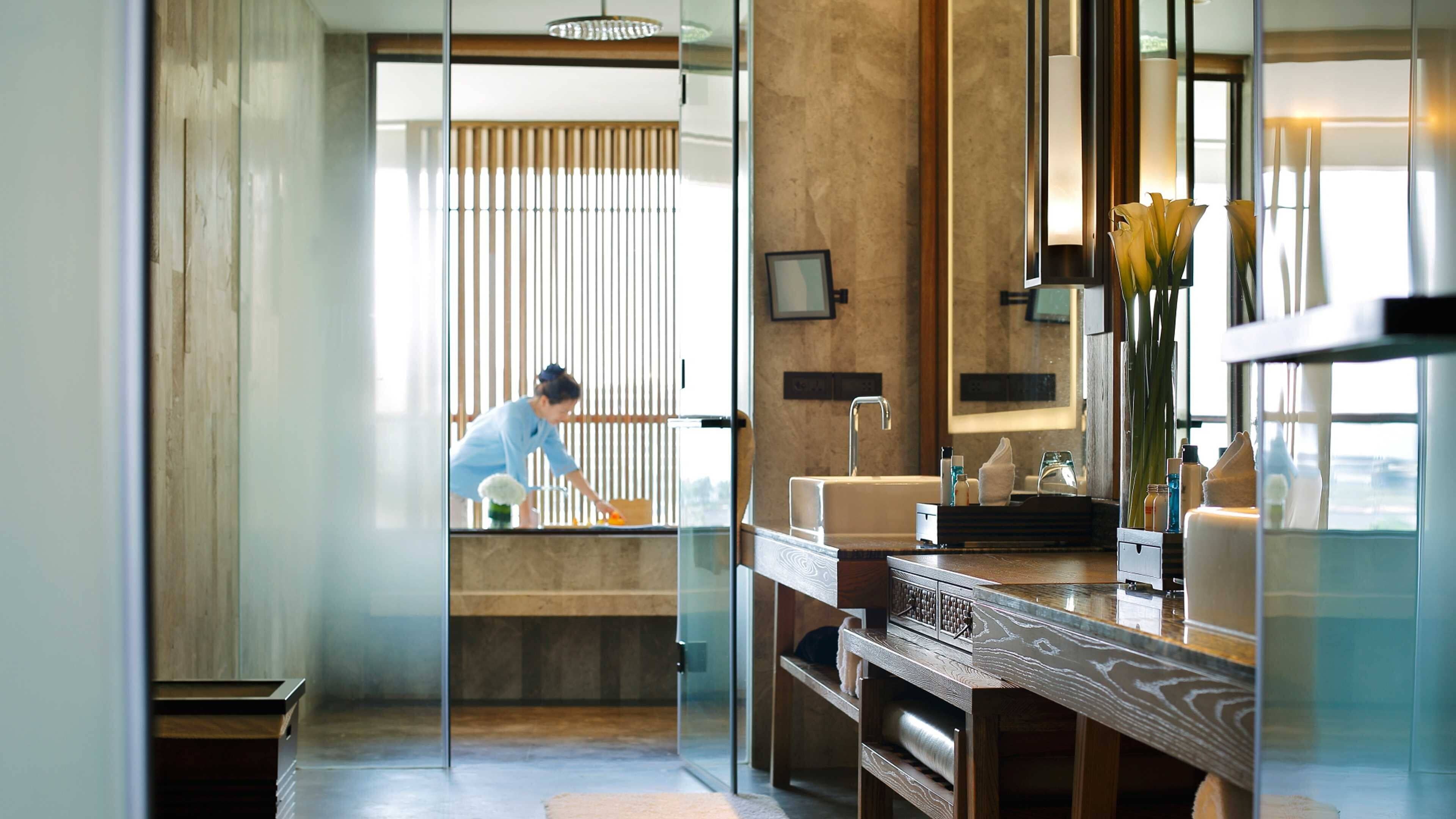 Un baño moderno con separador de vidrio, detalles de madera, un lavamanos con artículos de aseo y un espejo grande.