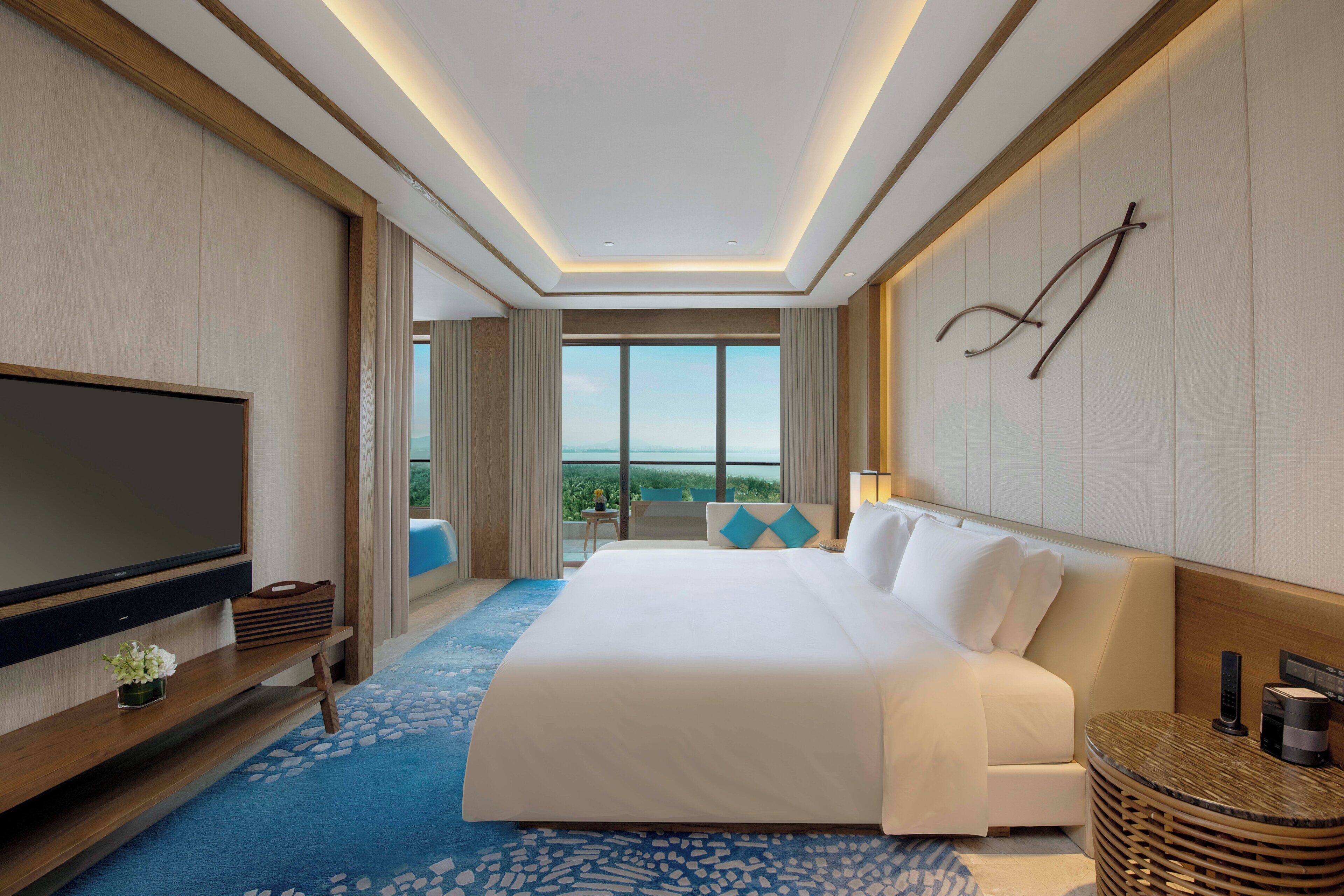 Habitación de hotel con una cama grande, un televisor de pantalla plana, una consola de madera, una mesita pequeña y vistas al exterior.