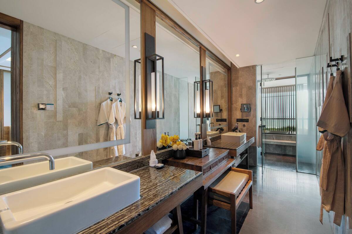 A modern bathroom with a large mirror, two sinks, a bench, and a glass shower enclosure.