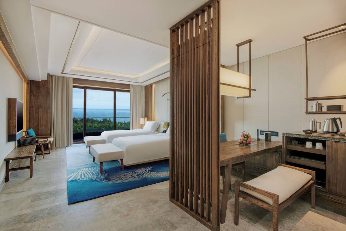 A hotel room with a large bed, two bedside tables, a desk, a chair, a view of the outdoors, and a kitchenette.