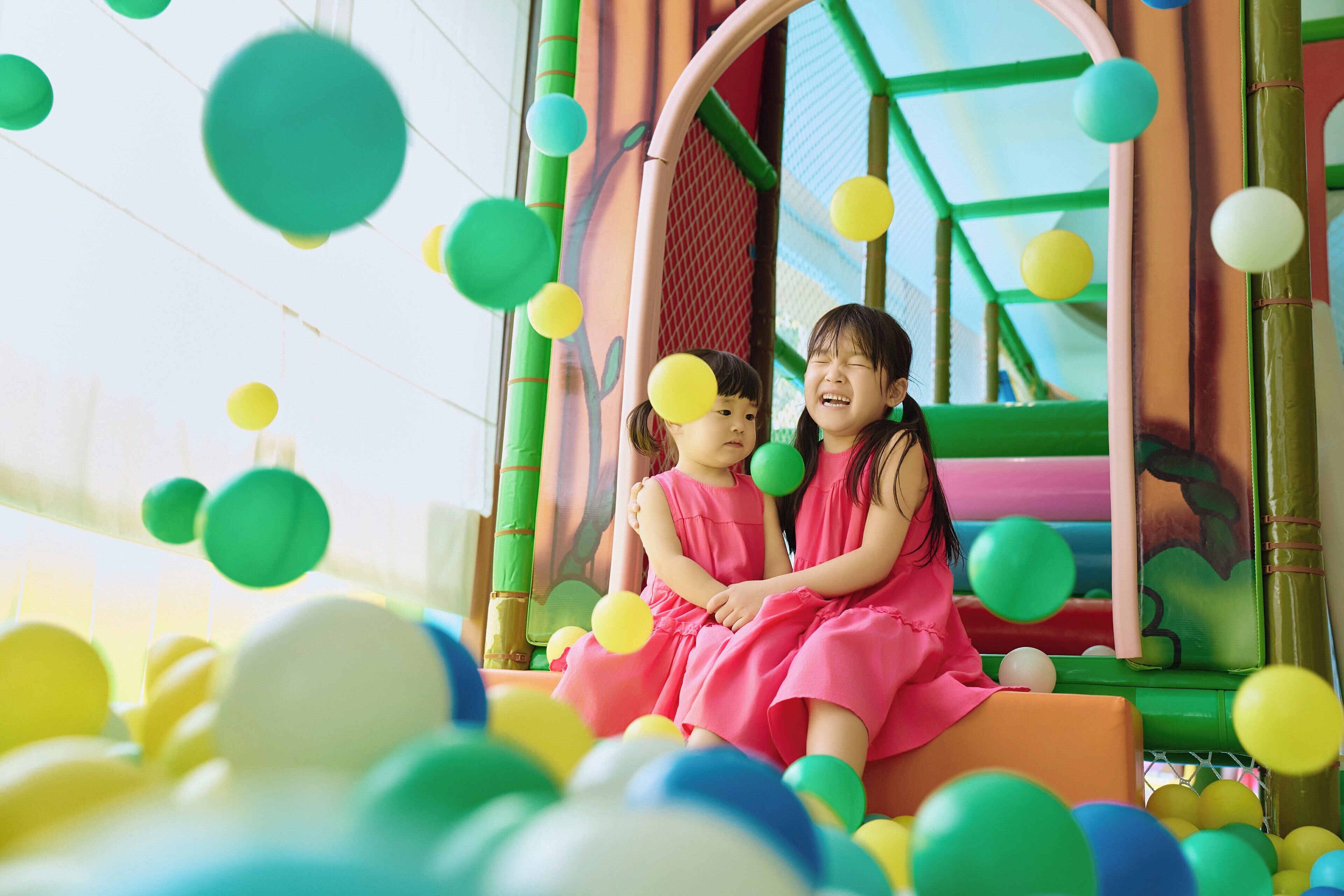 Two children in pink dresses sitting on a colorful play structure surrounded by a ball pit.
