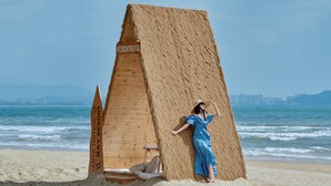 Eine Person steht neben einer großen Struktur aus Heuballen am Strand, im Hintergrund ist eine Stadtlandschaft zu sehen.