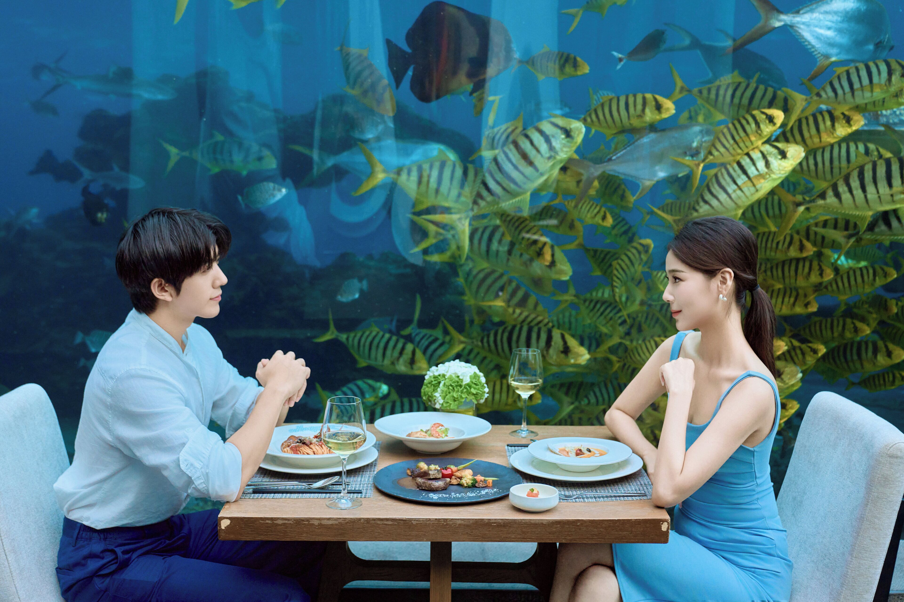 A dining table with plates of food and glasses, set against a backdrop of a large aquarium with various fish.