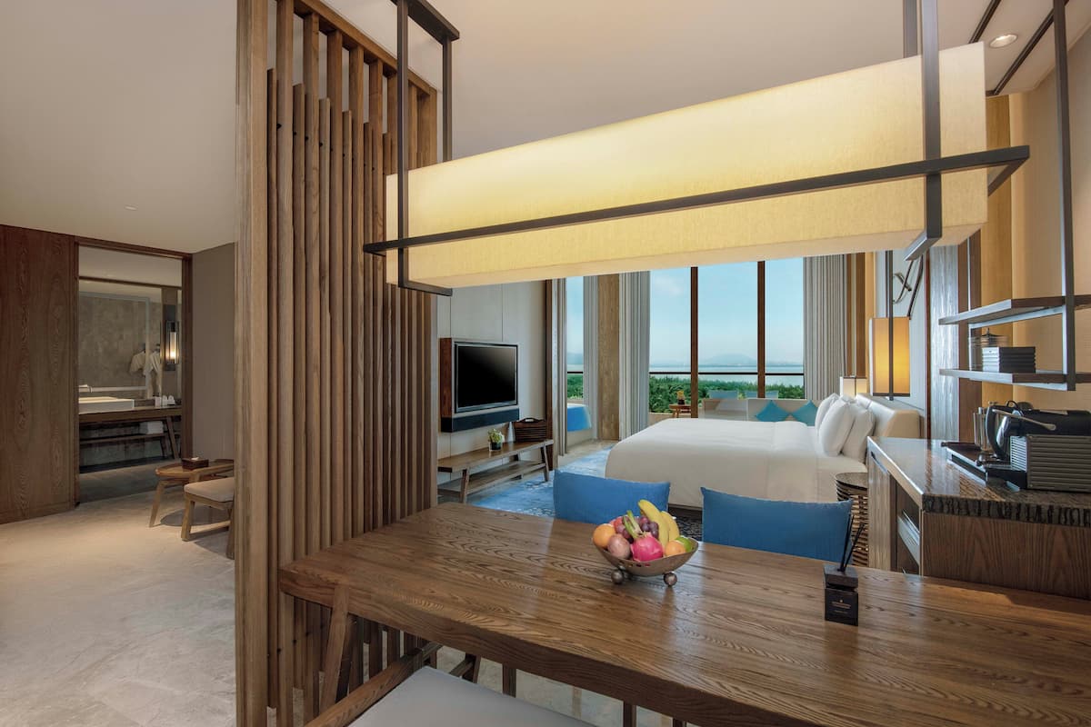 A modern hotel room with a wooden dining table, a fruit bowl, a bed with a city view, a TV, and a balcony.