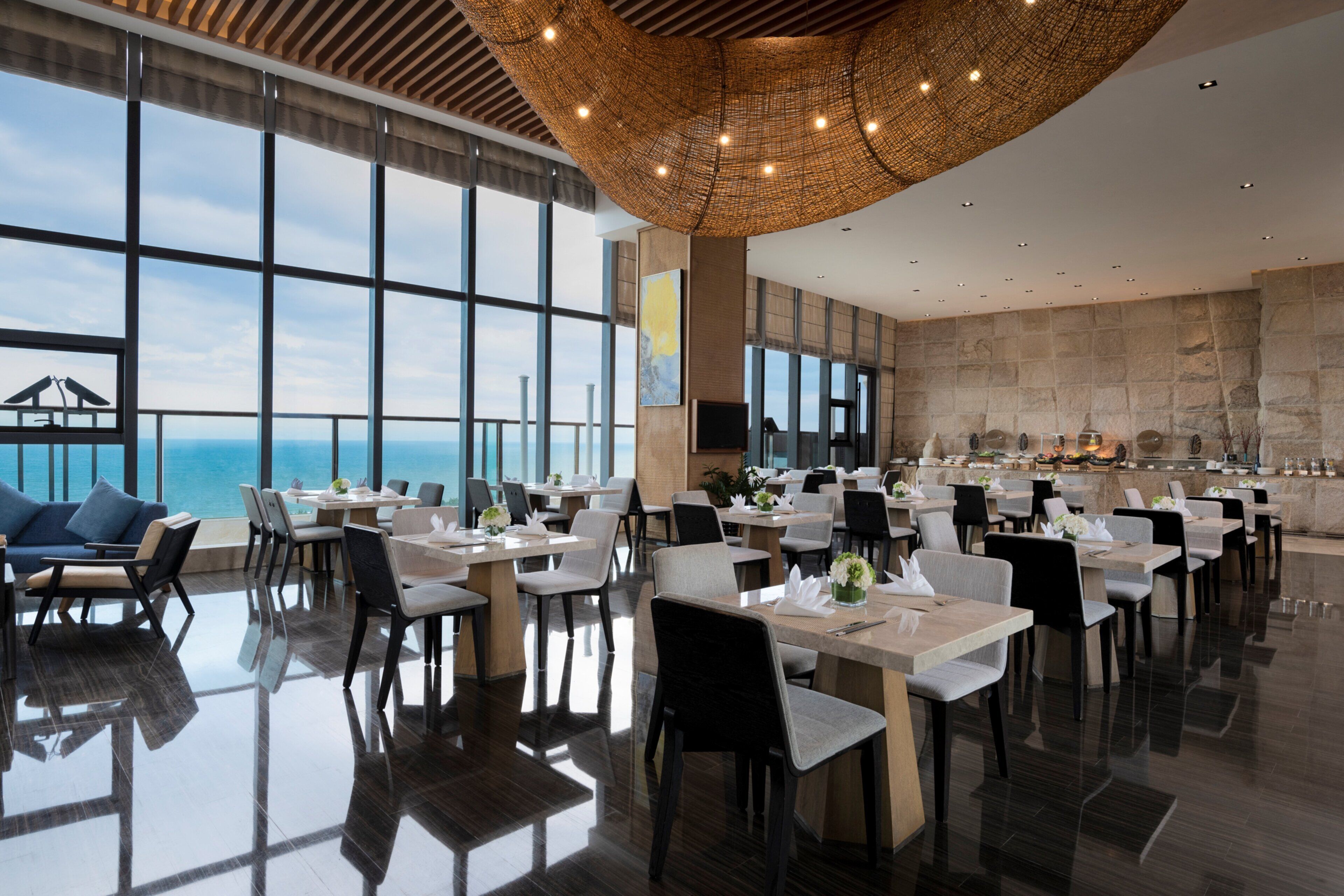 Un comedor amplio con mesas y sillas, ventanas grandes que ofrecen vistas al océano y un diseño de techo moderno.