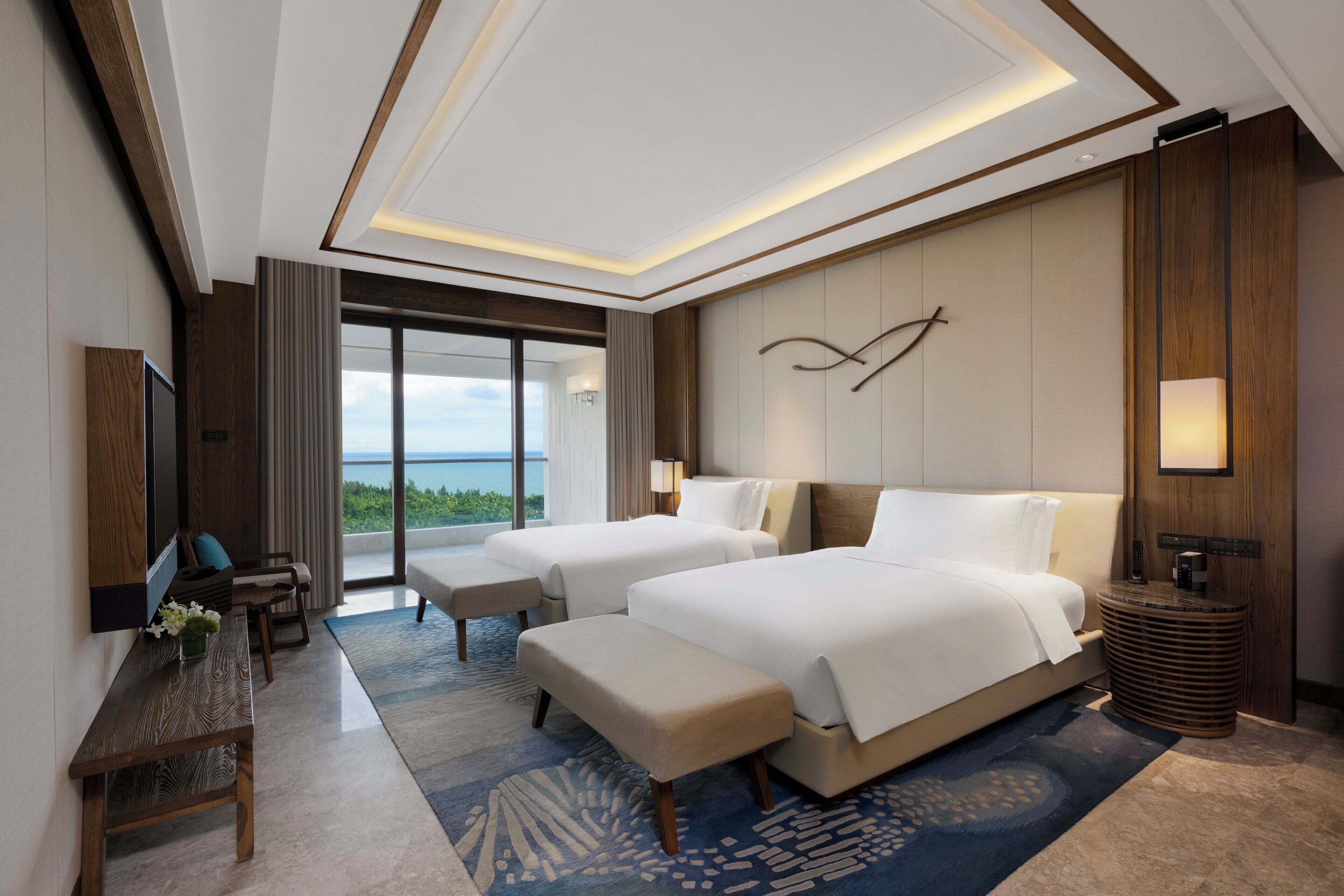 A modern hotel room with two beds, a desk, and a large window offering a view of the ocean.