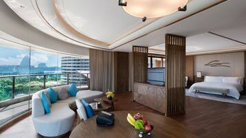 A modern hotel room with a curved ceiling, a sofa with blue cushions, a dining table with fruit, a bed, and a view of the city.