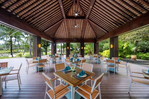 A covered outdoor dining area with wooden tables and chairs, a ceiling fan, and a view of tropical greenery.