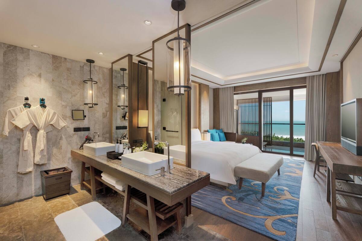 A modern hotel room with a large bed, a sitting area, a bathroom with a large mirror, and a view of the ocean.