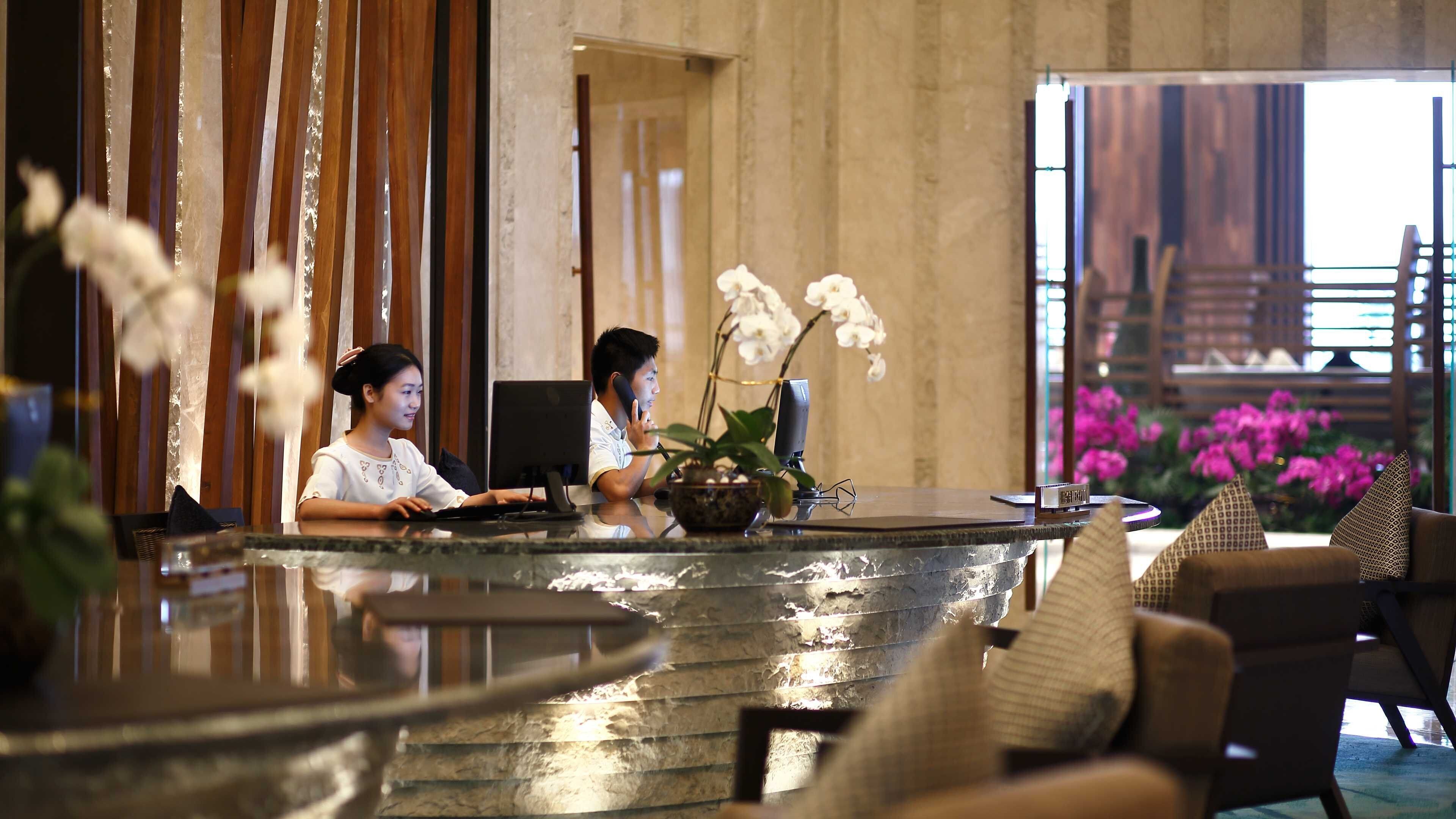 A reception desk with a computer, a plant, and a sign. There are chairs and a window with flowers visible.