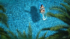 Eine Person sitzt auf einer schwimmenden Plattform in einem klaren, blauen Pool, umgeben von Palmen.