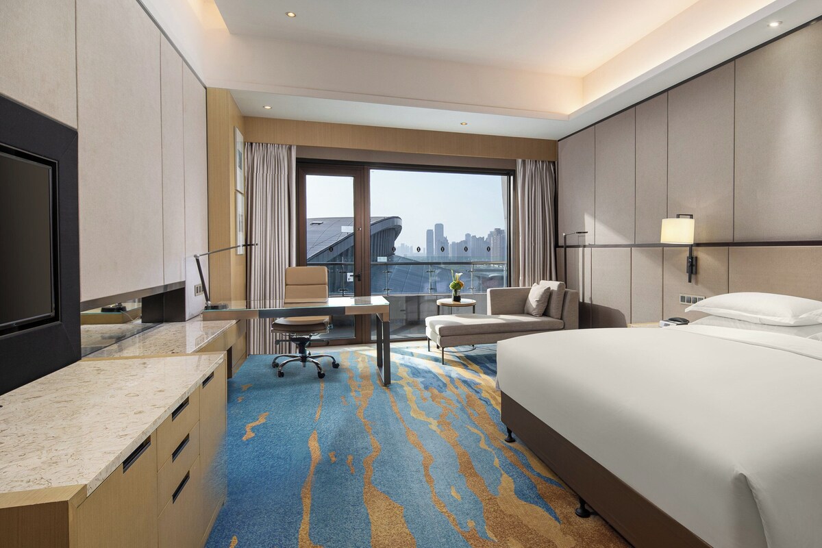 A hotel room with a large bed, a desk with a chair, a small table with a lamp, and a view of a cityscape through the window.