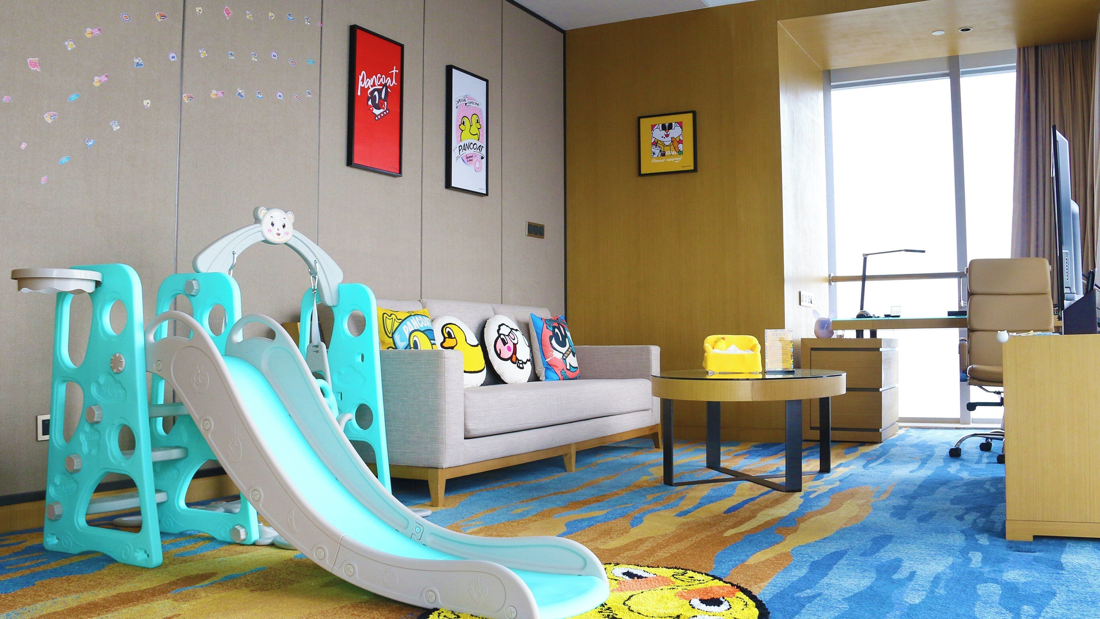 A hotel room with a sofa, a small table, a desk, a chair, a desk lamp, a window with curtains, a colorful carpet, and a children's slide.