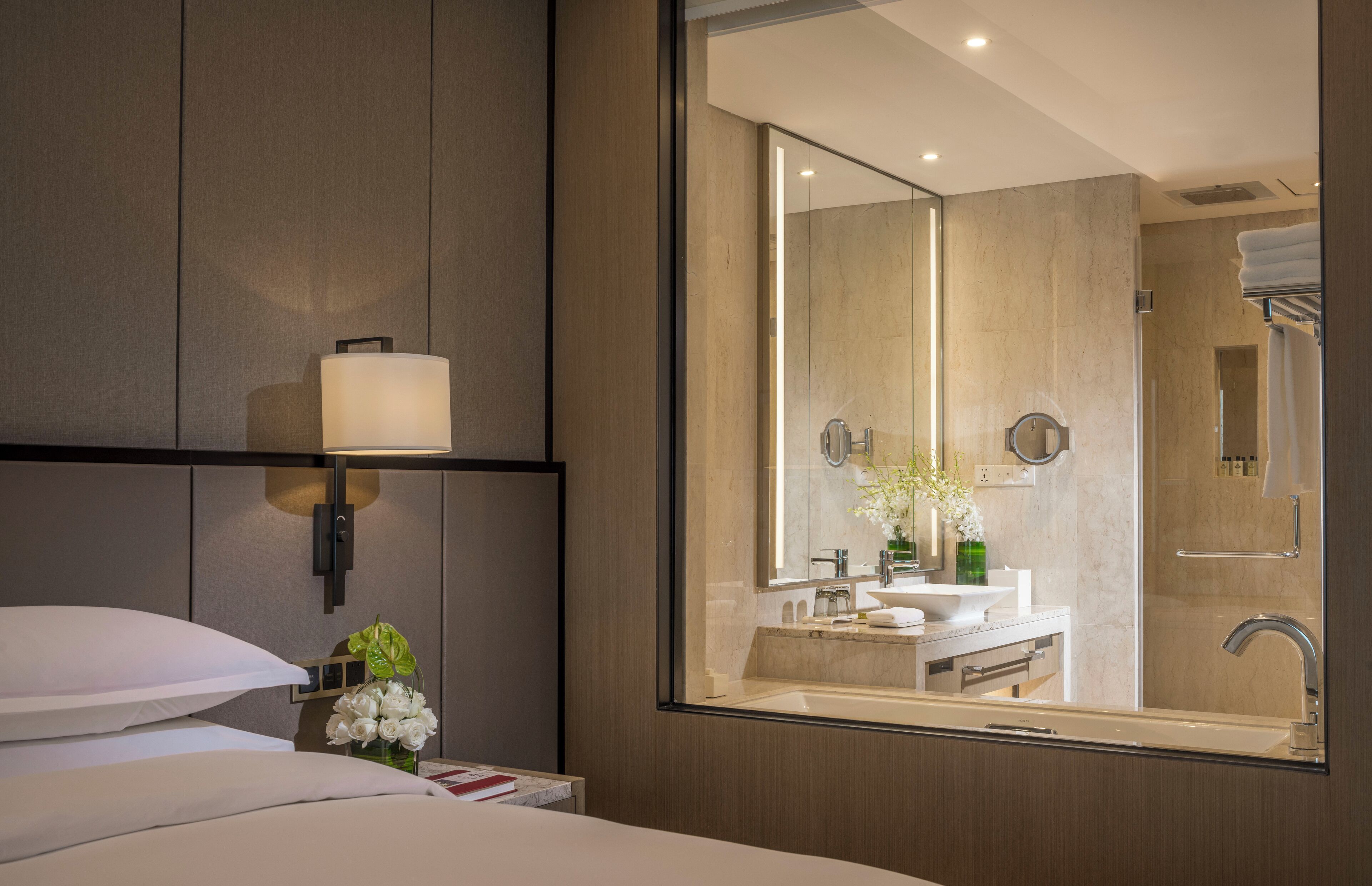 A modern hotel room with a large bed, a bedside lamp, a vase with flowers, and a view of the bathroom through a mirror.