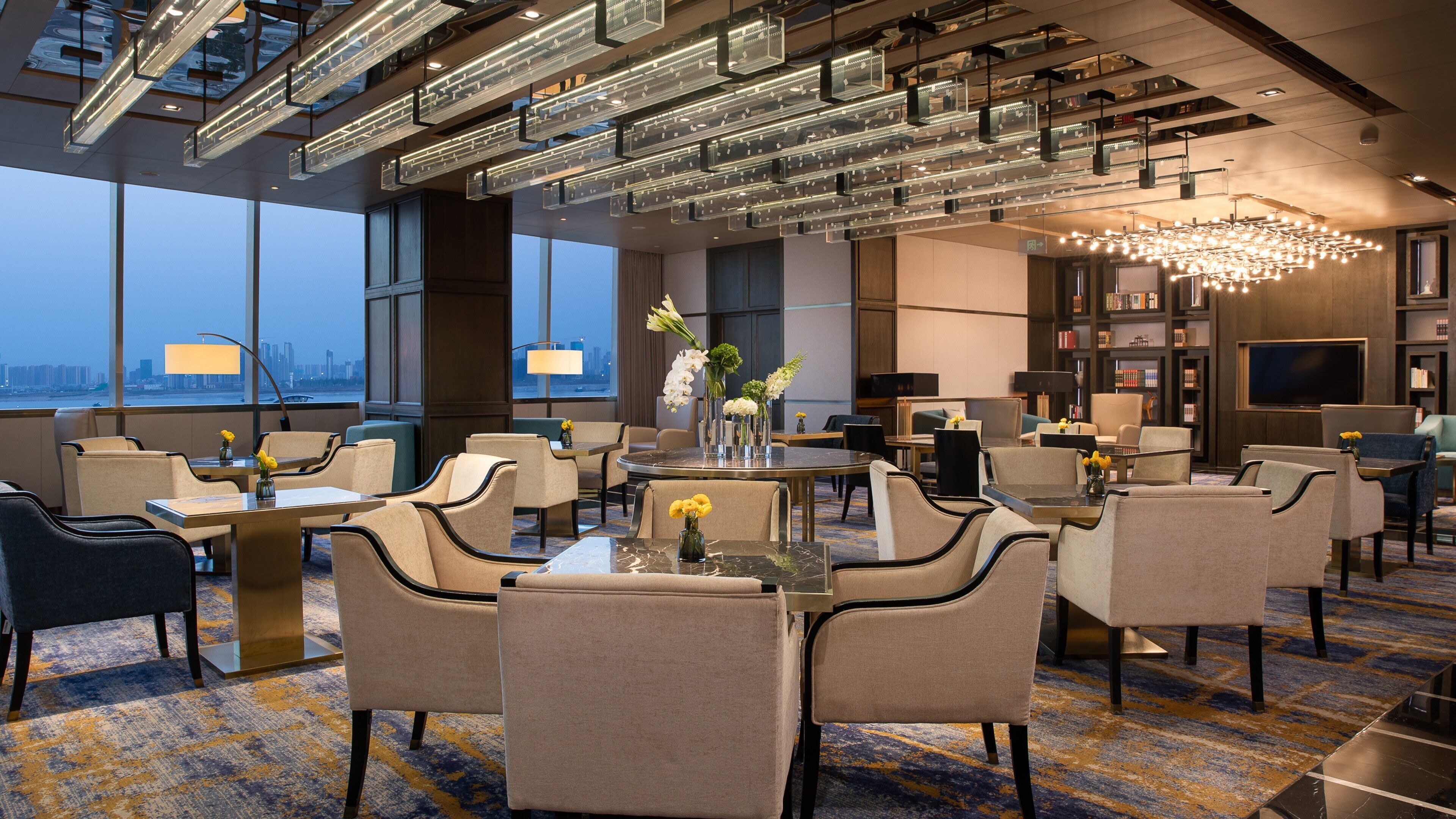 Un hall d’hôtel au design moderne, avec une grande table ronde entourée de chaises, un lustre et une bibliothèque remplie de livres.