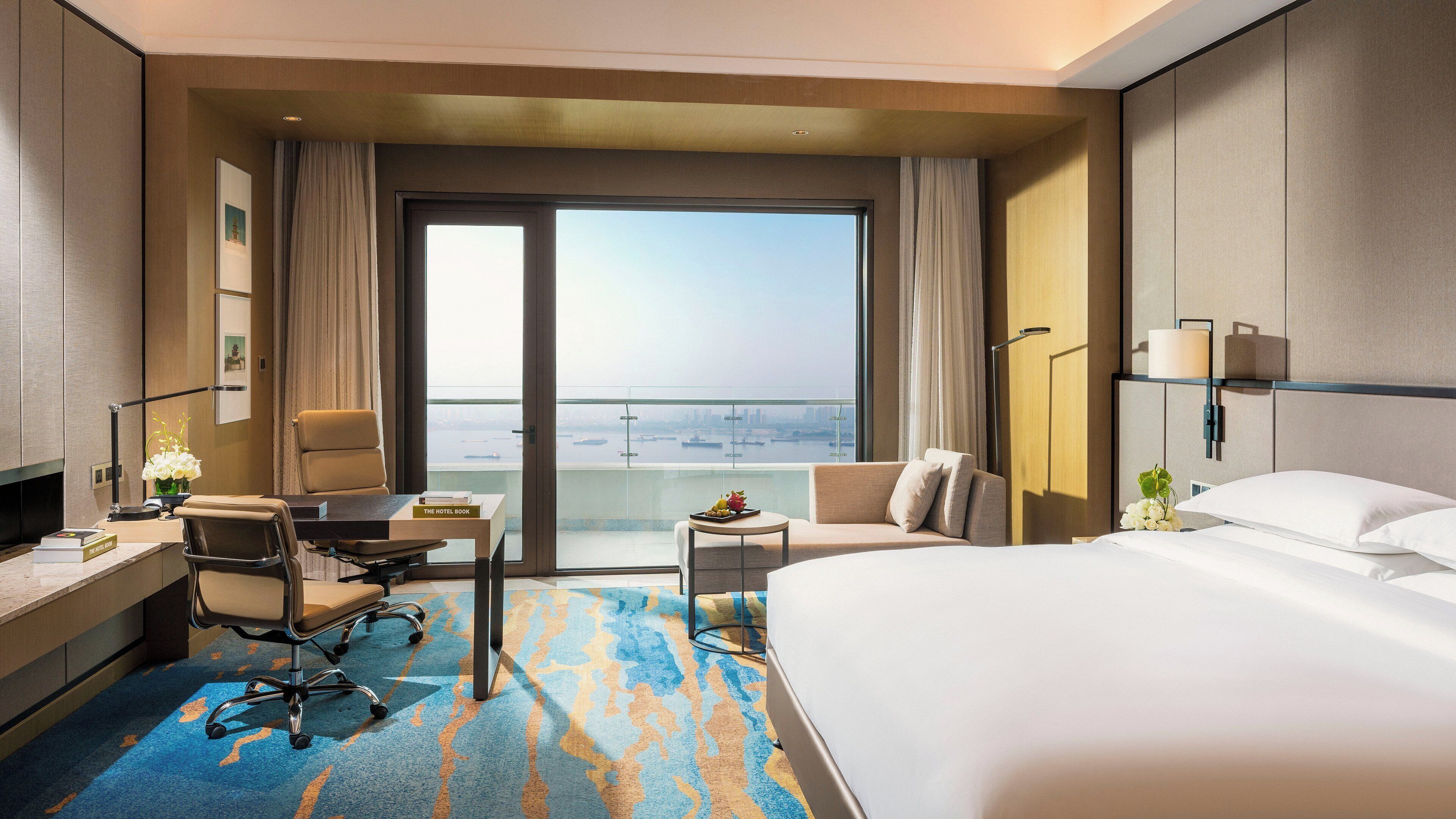 A hotel room with a bed, a desk with a chair, a small table with a fruit plate, a sofa, and a view of the city through a large window.