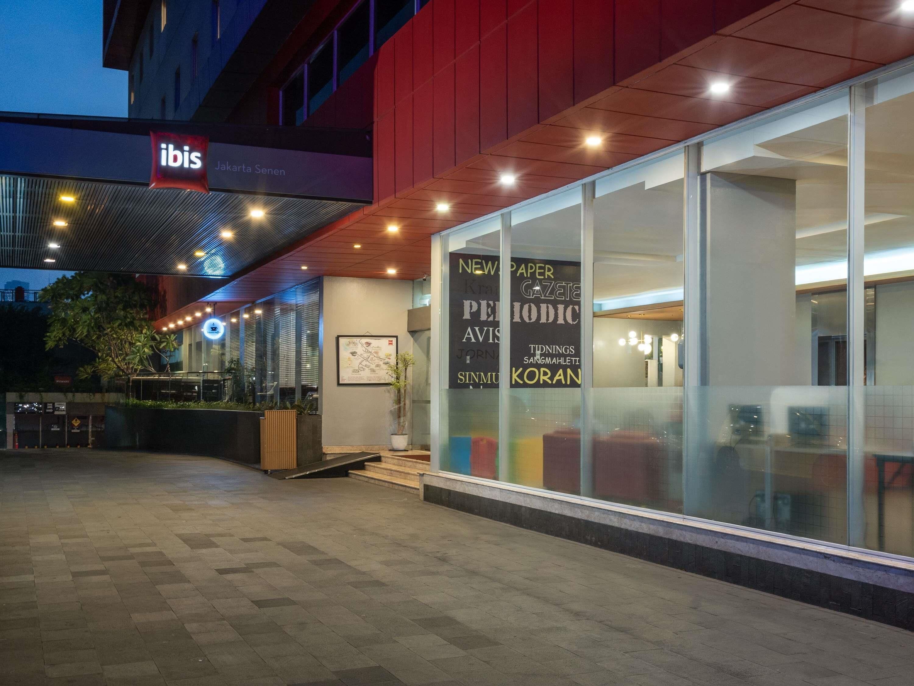 Un hôtel moderne avec une façade en verre, un mur extérieur rouge et une enseigne indiquant « ibis Jakarta Senen ».