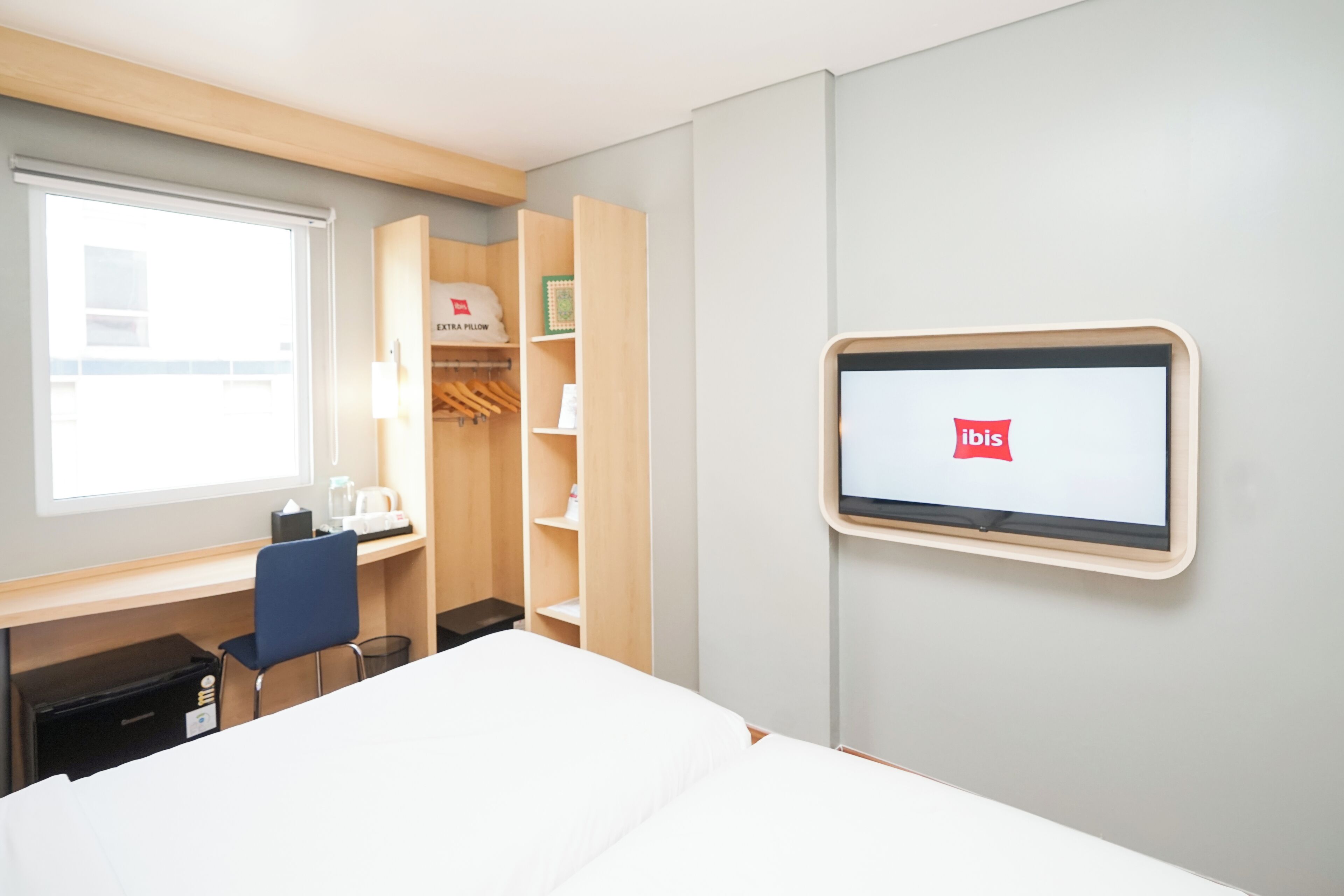A hotel room with a bed, a desk, a chair, a wardrobe, and a TV screen displaying the ibis logo.