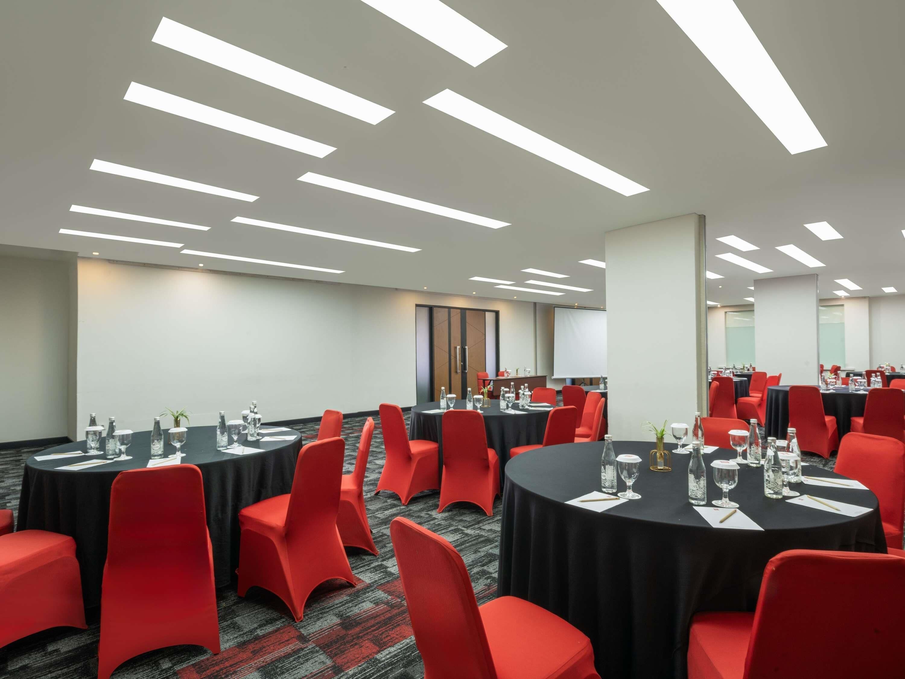 Un salón de conferencias con varias mesas, sillas rojas e iluminación colgante.