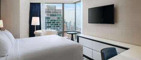 A hotel room with a large bed, a desk, a chair, a TV mounted on the wall, and a view of the cityscape through large windows.