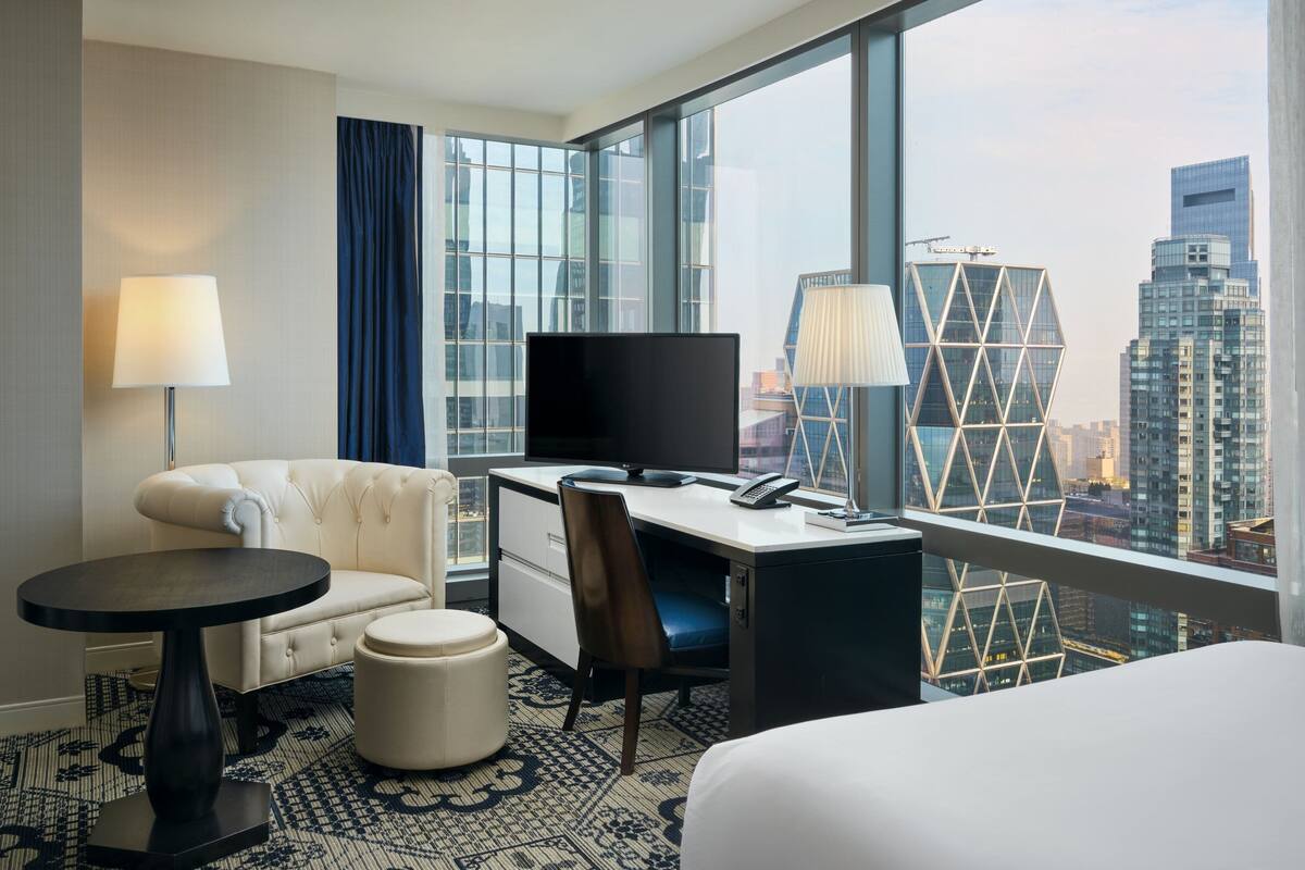 A hotel room with a large window, a bed, a desk with a computer, a chair, a small table, and a lamp.