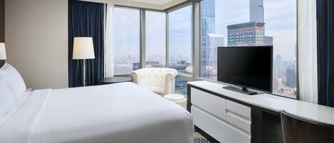 A hotel room with a large bed, a desk with a TV, a chair, and a view of a cityscape through large windows.