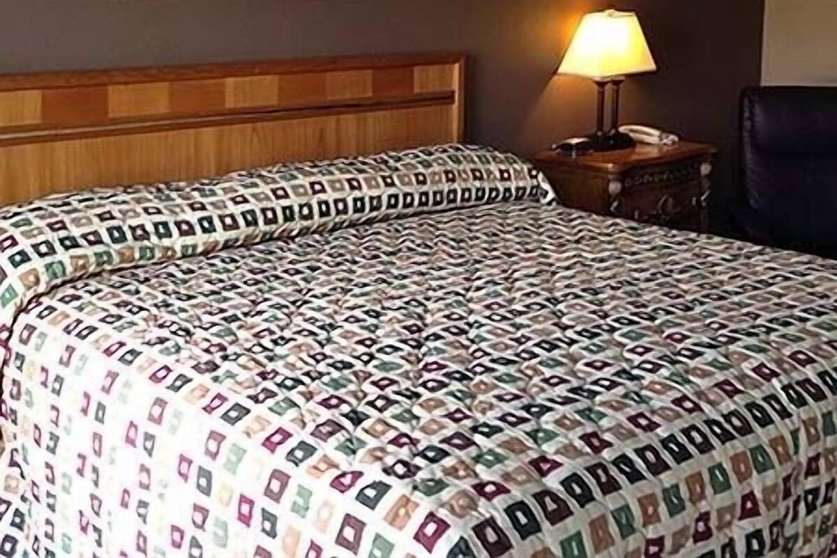 Una habitación de hotel con una colcha estampada, cabecero de madera, un cuadro enmarcado en la pared, una mesita de noche con lámpara y una silla negra.