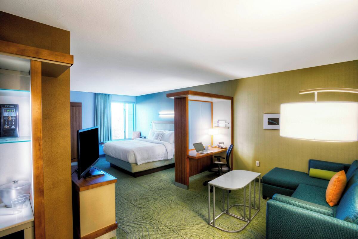 A hotel room with a bed, a desk with a laptop, a TV, a sofa, and a small table.