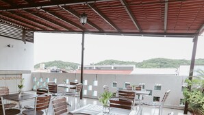 Eine Terrasse im Freien mit Tischen und Stühlen, gefliestem Boden und Blick auf ein Gebäude sowie Grünpflanzen im Hintergrund.