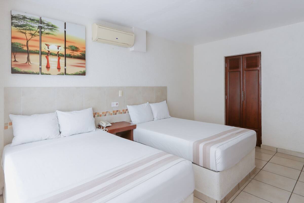 Habitación de hotel con dos camas, un armario de madera, aire acondicionado de pared y un cuadro de una puesta de sol sobre una sabana.