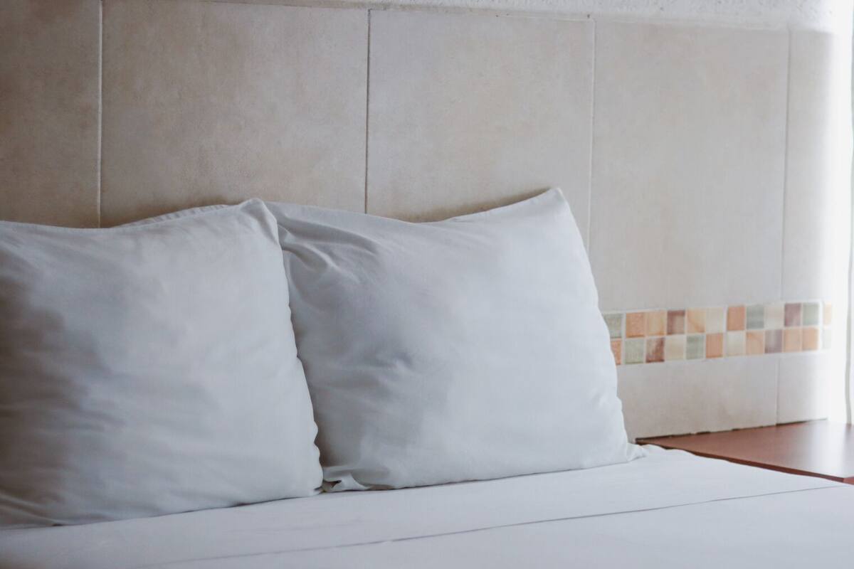 Una cama bien hecha con sábanas blancas y dos almohadas, frente a una pared con zócalo de azulejos.