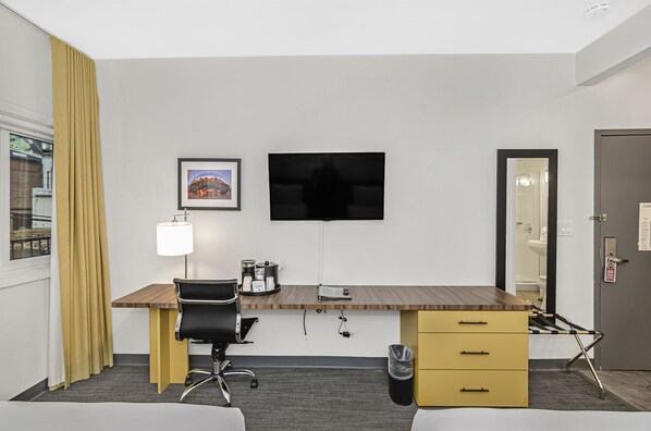 Habitación de hotel con escritorio de madera, silla de oficina negra, televisor montado en la pared, un pequeño cubo de basura y un mueble de almacenamiento amarillo.