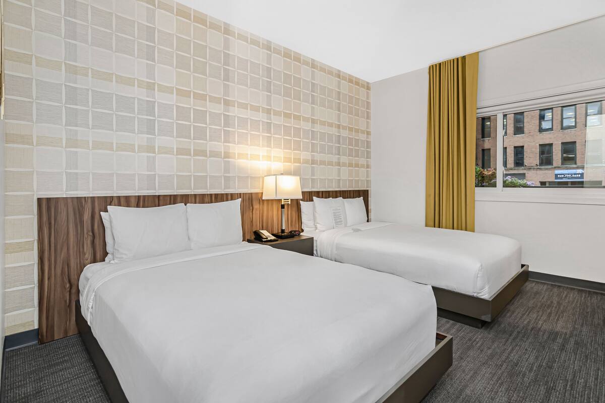 A hotel room with two beds, a wooden headboard, a nightstand with a lamp, and a window with a view of buildings.