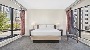 Habitación de hotel con una cama grande, dos mesitas de noche con lámparas, una silla y vistas a la ciudad a través de ventanas amplias.