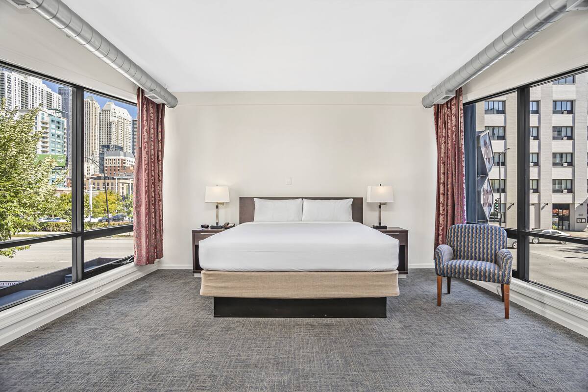 A hotel room with a large bed, two bedside tables with lamps, a chair, and a city view through large windows.