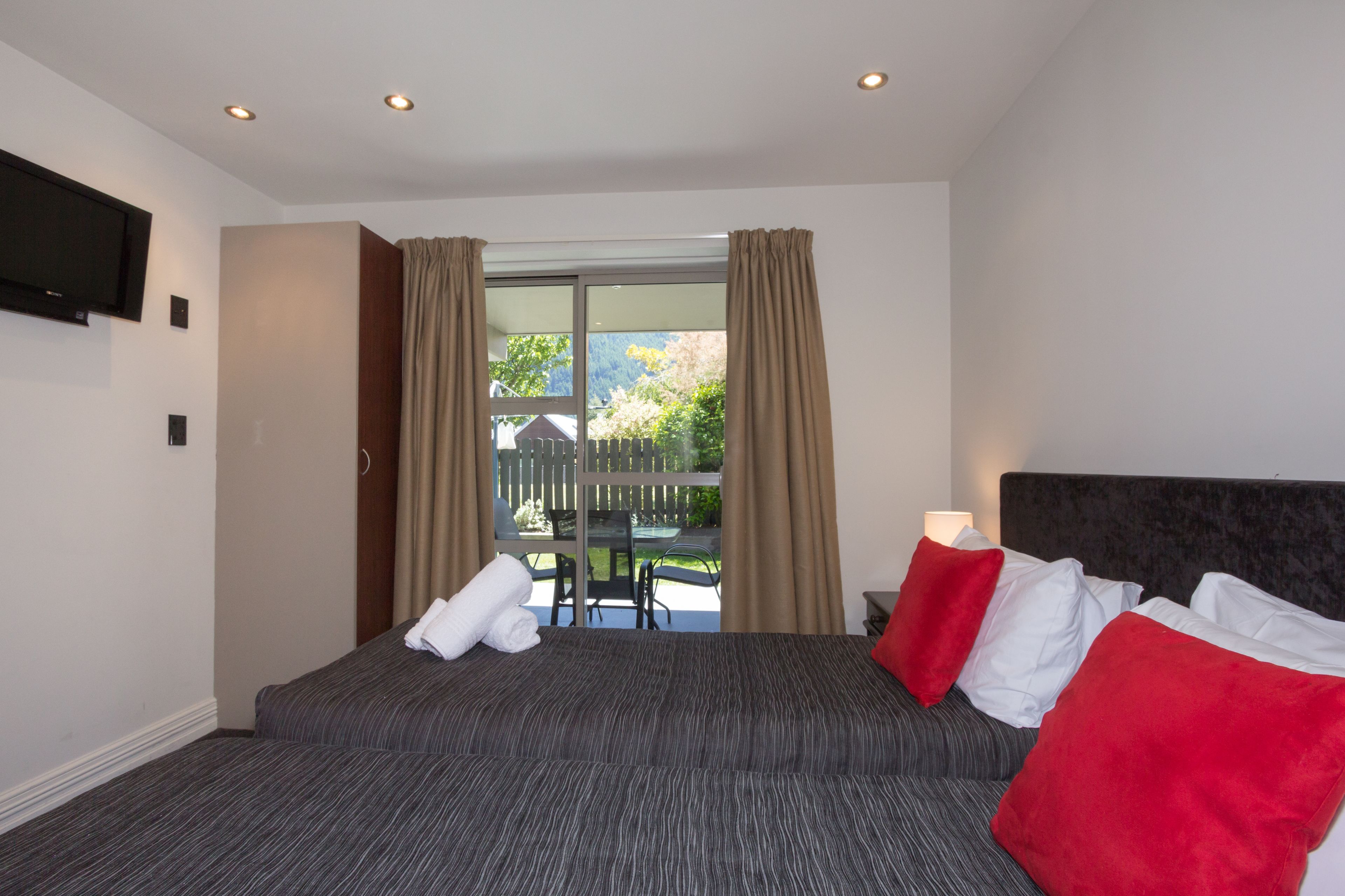 A hotel room with a bed, a TV mounted on the wall, and a sliding glass door leading to an outdoor area with a table and chairs.