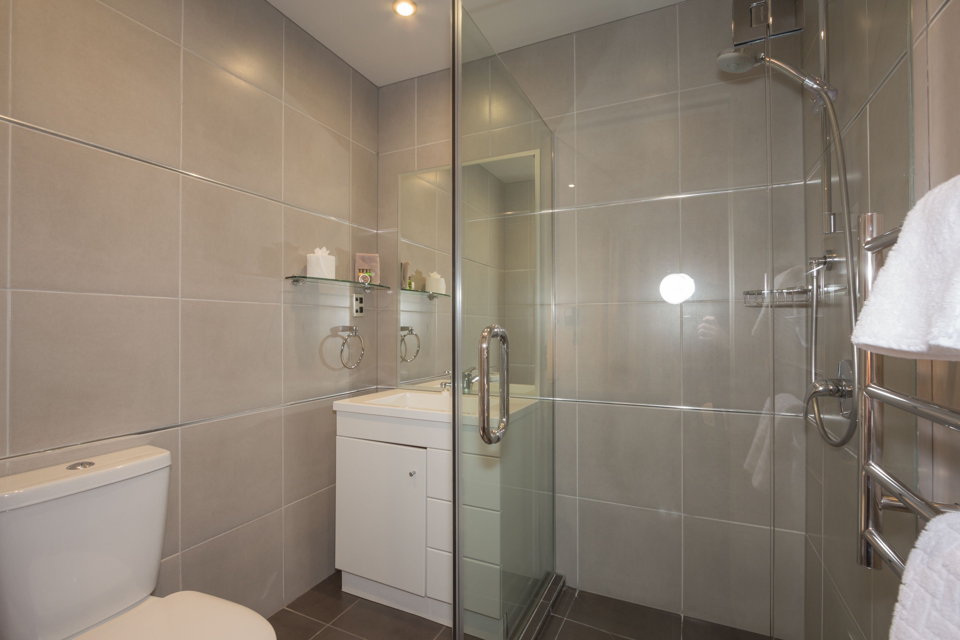 A modern bathroom with a glass-enclosed shower, a white toilet, and a glass-fronted sink with shelves above.