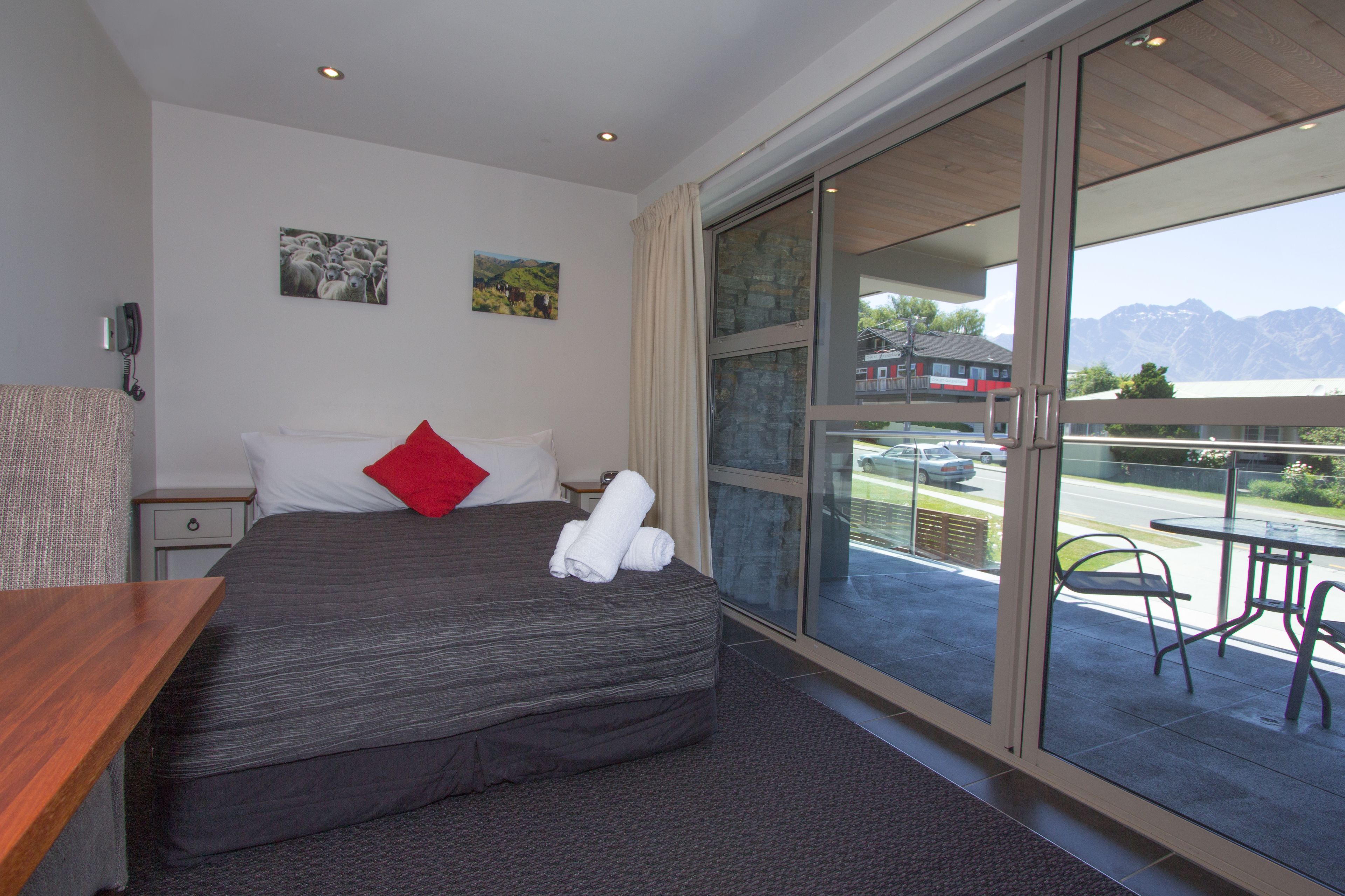 A hotel room with a large bed, a wooden desk, a small bedside table, and a view of the mountains through a large window.