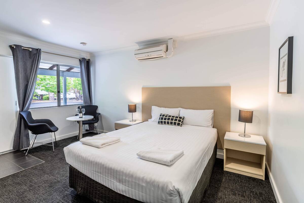 Une chambre d’hôtel avec un grand lit, deux tables de chevet avec des lampes, une petite table avec une chaise et une fenêtre avec des rideaux.