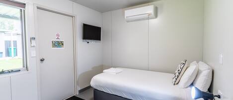 A small, clean room with a bed, a TV, an air conditioner, and a window.