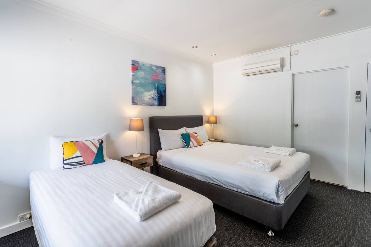 Une chambre d’hôtel avec deux lits, chacun muni d’un oreiller et d’une lampe de chevet. Un tableau est accroché au mur et un appareil de climatisation est installé au-dessus de la porte.