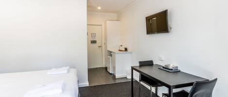 Quarto de hotel com cama, escrivaninha e cadeira. Há uma televisão na parede e uma kitchenette pequena ao fundo.