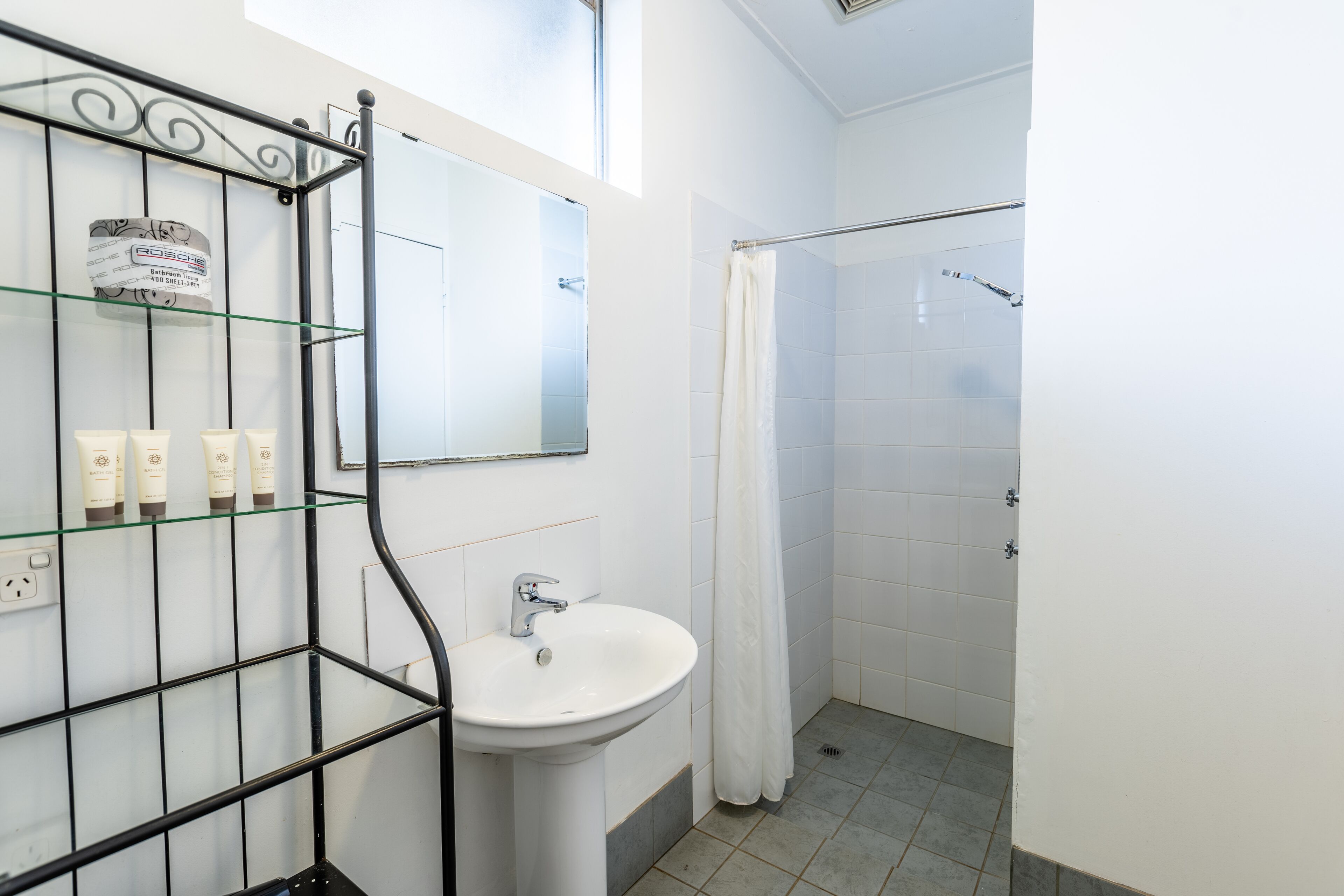 A bathroom with a glass shelf holding toiletries, a white sink, and a shower with a curtain.
