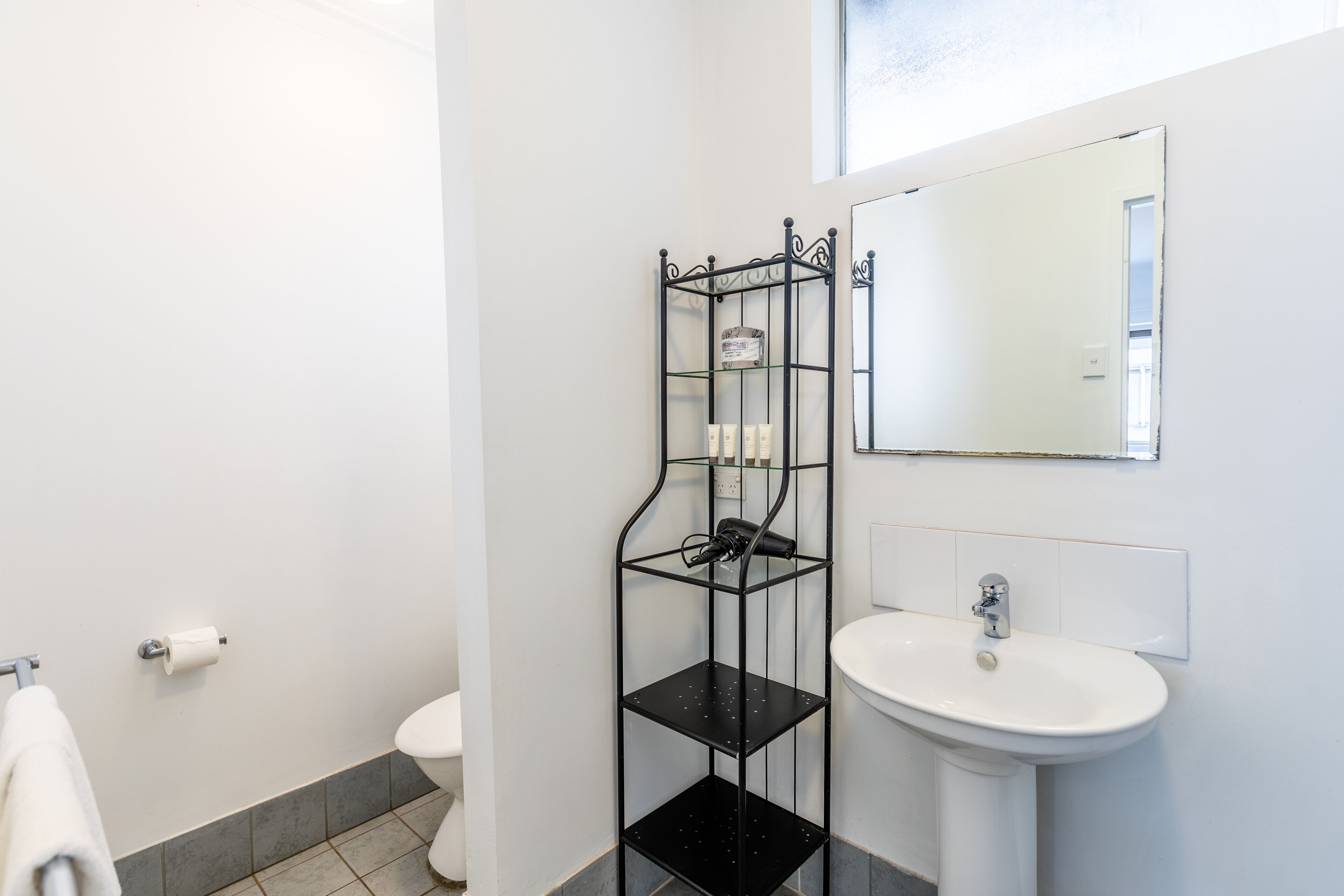 Un baño con un lavamanos blanco, un estante de metal negro con artículos de aseo, un inodoro y un perchero para toallas.